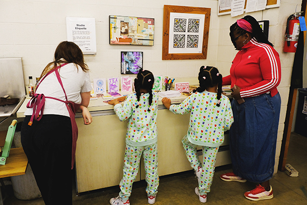 An artist wearing an apron demonstrates printmaking to two kids in matching outfits and their mom.