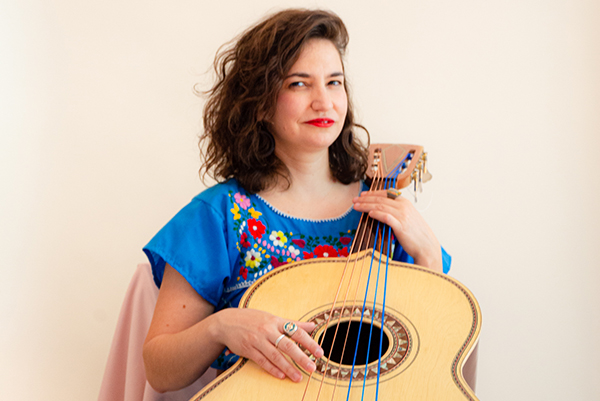 A woman in a blue top with colorful flowers embroidered on it holds an acoustic guitar. Three of the guitar strings are also blue.