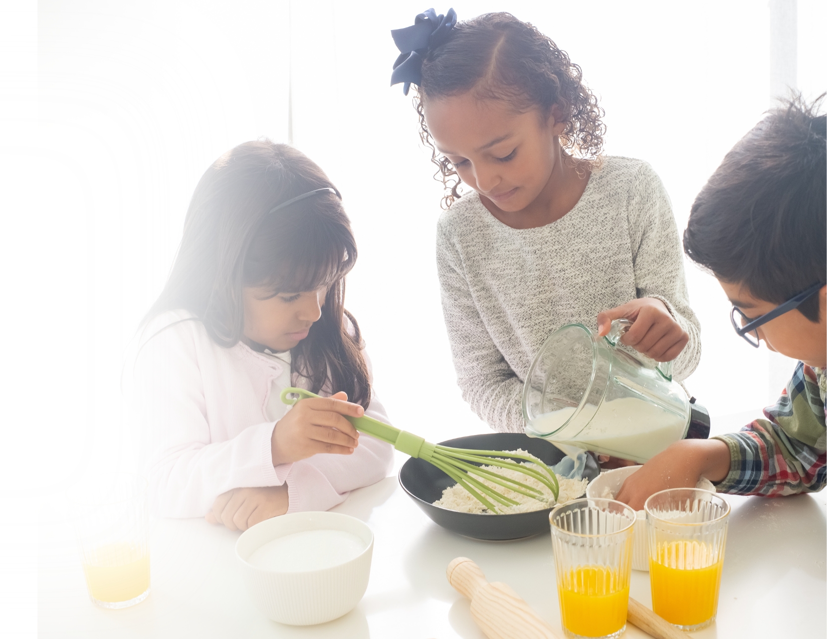 Three young children work together at a white table mixing ingredients in a bowl and pouring milk from a blender.