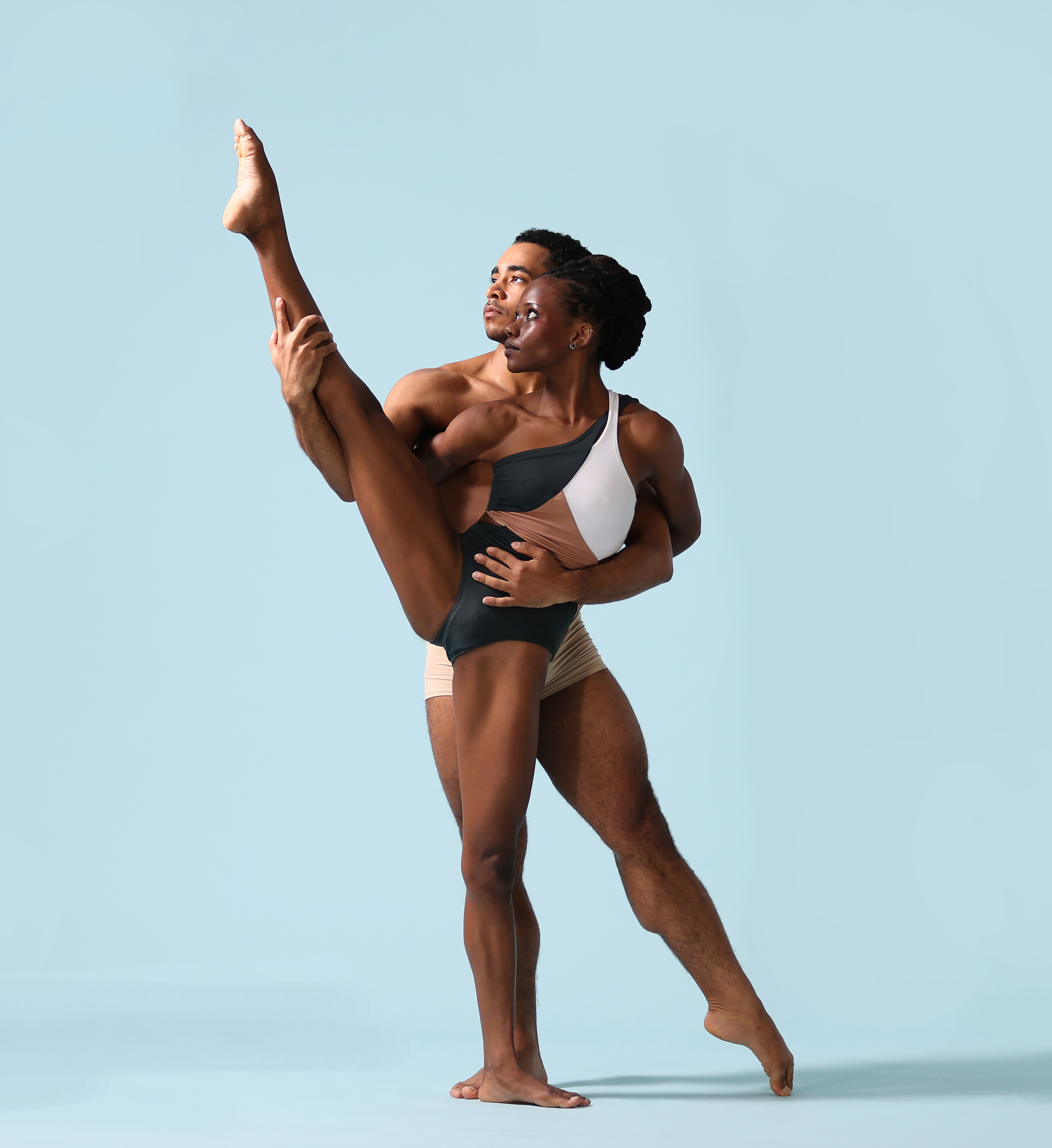Two dancers, a man and a woman, pose in a supportive partner stretch against a light blue background.