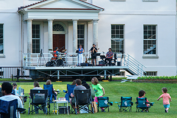 Musicians perform on an outdoor stage in front of a historic building as families watch from lawn chairs.